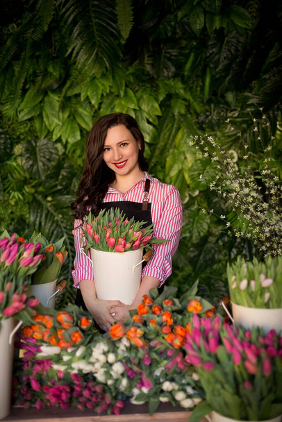 Portrait d'une fleuriste au travail dans son atelier avec décor de feuilles tropicales. Une belle fille dans un tablier de travail se tient derrière le comptoir d'un magasin de fleurs et fait des bouquets avec des tulipes colorées. - Photo, image