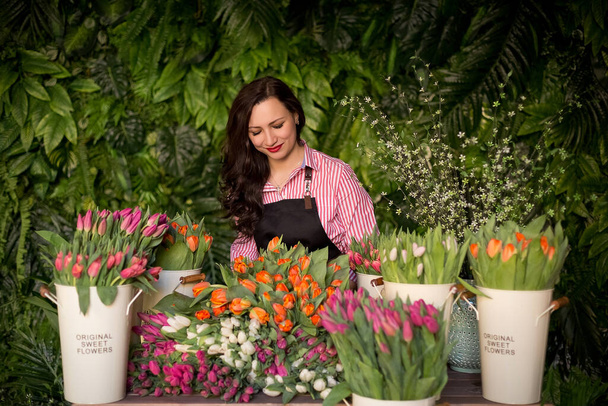 Muotokuva kukkakauppias töissä hänen studio trooppisia lehtiä sisustus. Kaunis tyttö työssä esiliina seisoo tiskin takana kukkakaupan ja tekee kukkakimppuja värikkäitä tulppaanit. - Valokuva, kuva