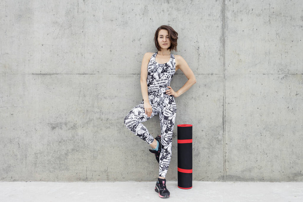 Fitness girl in black and white fitness suite with gym mats outdoor. Against a gray concrete wall. - Photo, Image