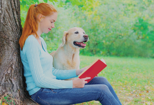 Young woman reading a book with Golden Retriever dog sitting near a tree in a park - Foto, immagini
