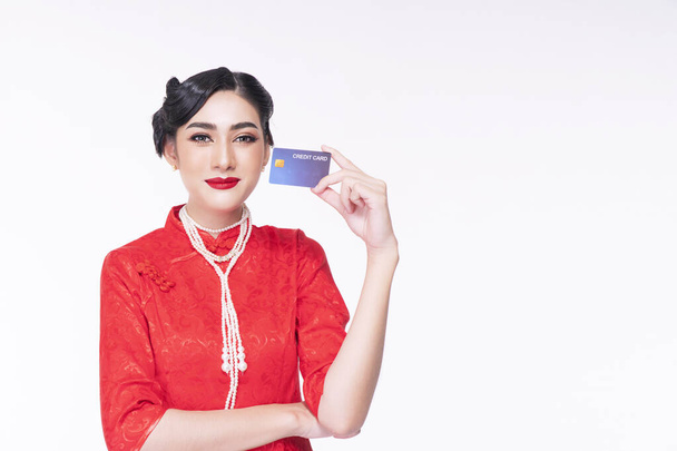 Confiance dans le concept de paiement. Jeune femme porter rouge cheongsam robe chinoise tenant la carte de crédit maquette tout en se tenant sur fond blanc isolé.  - Photo, image