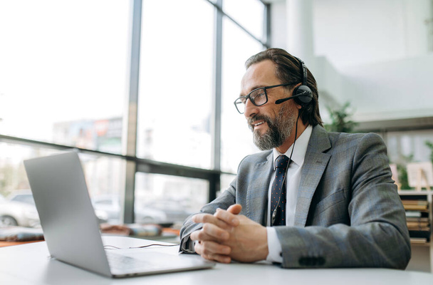 Mature male manager in headset and formal suit sits at the desk, work as call center operator, communicate with clients. Caucasian businessman is consulting people, support service concept - Photo, Image