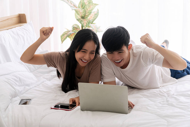Opgewonden Aziatisch paar kijken spel of winnaar bieden of tv online of examen met laptop terwijl ze samen op het bed thuis liggen. Lifestyle internet technologie en nieuw normaal concept. - Foto, afbeelding