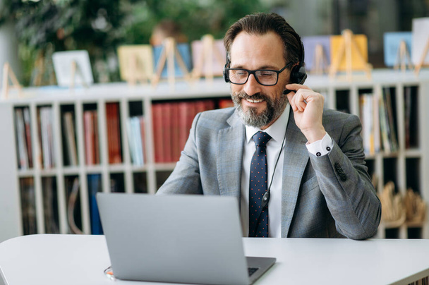 Caucasian male employee in headphones uses laptop, sitting at the desk. Senior satisfied businessman is consulting clients, smiling, work in support service as call center operator - Foto, Bild