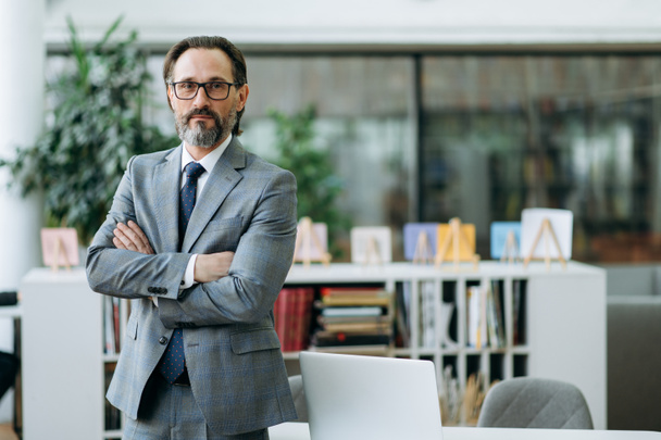 Portrait of successful mature caucasian businessman in formal suit and eyeglasses. Confident influential male entrepreneur stands with arms crossed in modern office, looks directly at camera - Фото, изображение