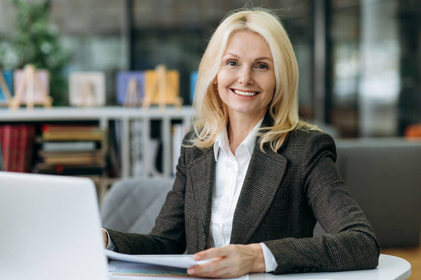 Πορτρέτο της ευτυχούς επιτυχημένης επιχειρηματία σε επίσημη ένδυση στο χώρο εργασίας. Κομψή ώριμη γυναίκα υπάλληλος κάθεται στο τραπέζι στο γραφείο, κοιτάζει την κάμερα με φιλικό χαμόγελο - Φωτογραφία, εικόνα