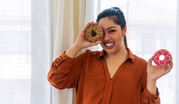 Snoepgoed junk food voor ongezond concept. Gelukkig glimlach obesitas dikke vrouw met chocolade chips koekjes bedekken haar ogen en donut in de hand, terwijl staan in de kamer thuis. - Foto, afbeelding