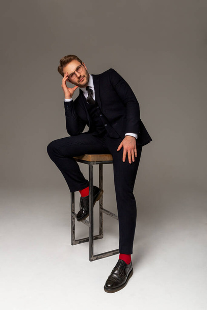 A man in a dark suit, white shirt, tie, black boots and glasses sits on a high chair. Confident look. Business clothes. Portrait of a man. Success. - Photo, Image
