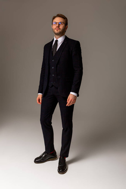 A man in a dark suit, white shirt, black boots and glasses is standing against a plain background, looking straight. Business clothes. Confident look. Success. Vertical photo - Foto, immagini
