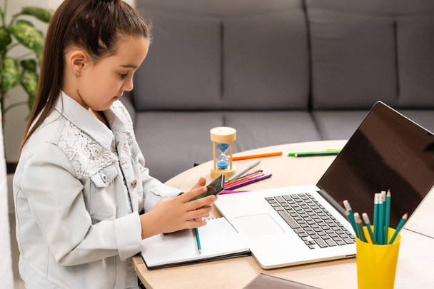 klein meisje zit met een laptop aan tafel en houdt een zandloper, wachten op het einde van online lessen - Foto, afbeelding
