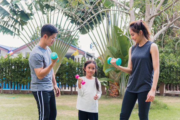 Asian young mother, father and child daughter doing exercising together with dumbbells is fun outdoors in nature a field garden park. Happy family kid sport and exercises for healthy lifestyle - 写真・画像