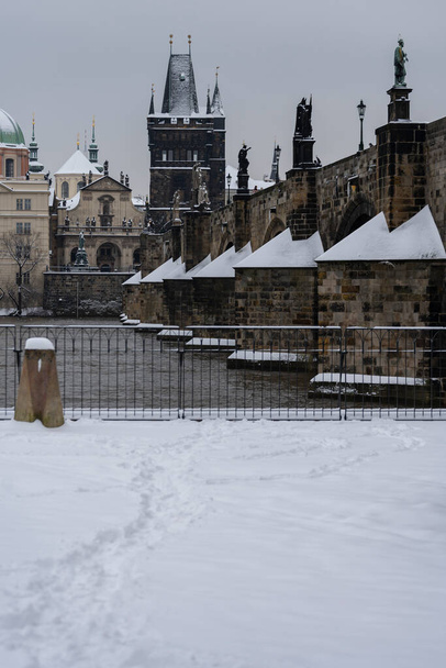 view of the snow-covered pillars of Charles Bridge on the Vltava River, snow on the roofs of buildings and towers and on the sidewalk in the center of old Prague - Photo, Image