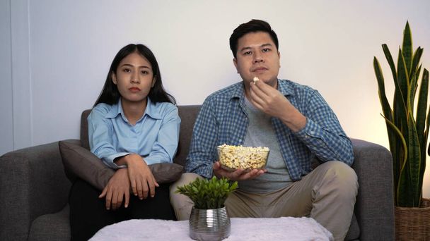 Feliz joven pareja asiática que pone sofá en su acogedora sala de estar viendo películas en la televisión y comiendo palomitas de maíz juntos en la noche en casa - Foto, imagen