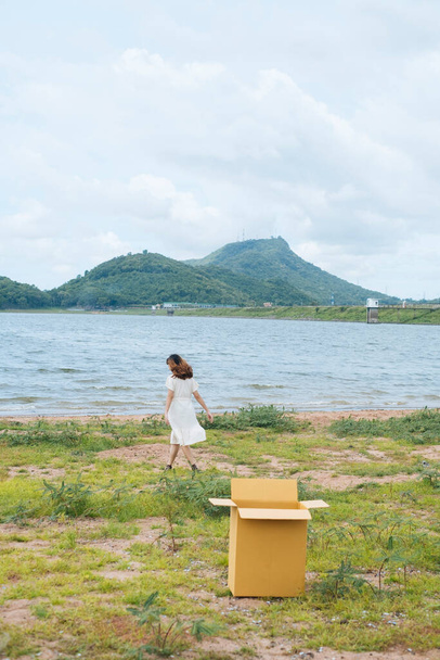 jong aziatische thai vrouw hipster krullend kapsel op wit jurk hebben plezier bij meer verlaten kartonnen doos achter. - Foto, afbeelding
