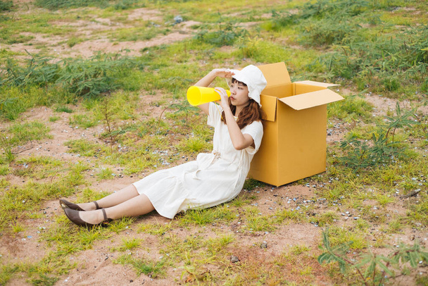 Jeune asiatique thai femme hipster bouclé coiffure sur robe blanche assis appuyé sur carton boîte et jouer avec jouet jaune parlant mégaphone. - Photo, image