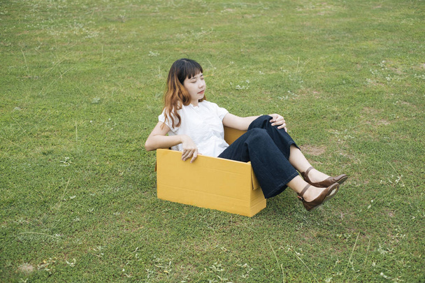 Fiatal ázsiai thai nő hipster göndör frizura fehér ing ül kartondobozon zöld park rét mező. - Fotó, kép