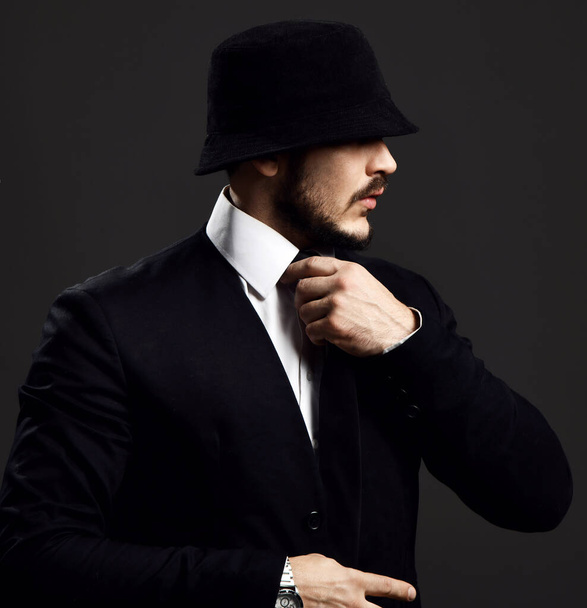 Young brutal bearded man in black suit and fashionable hat covering his eyes adjusts his tie looking aside - Photo, image