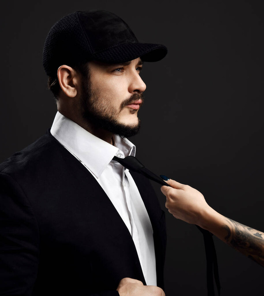 Female hand with tattoo is pulling young brutal bearded brunet man in fashionable cap and classical black suit by tie - Фото, зображення
