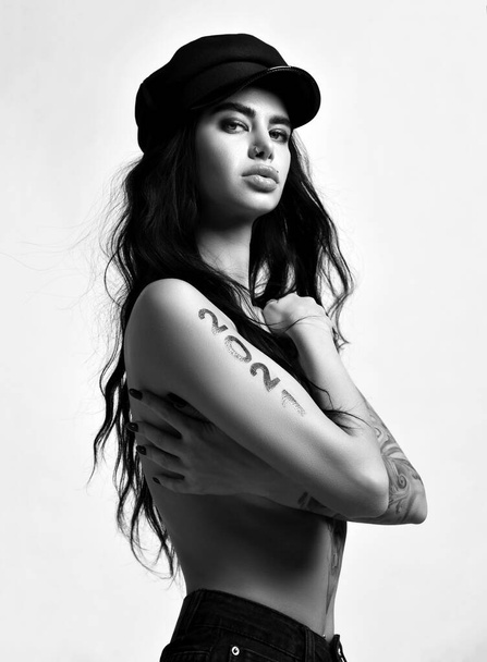 Black and white portrait of naked brunette woman with pouty lips piercing and tattoo 2021 on arms standing sideways - Valokuva, kuva