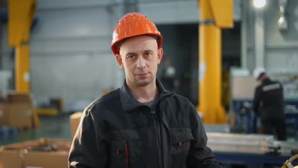 Un trabajador que lleva un casco en una fábrica mira atentamente a la cámara. Luego sonríe.. - Metraje, vídeo