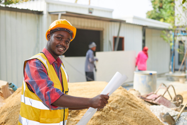 Een zwarte man draagt een veiligheidspak en een harde hoed met een papier in de hand, en hij kijkt naar de camera op een bouwplaats. - Foto, afbeelding