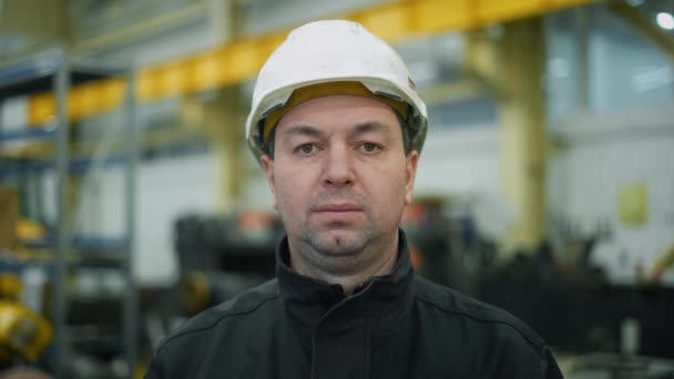 Een monteur in een helm staat in de fabriek en kijkt aandachtig in de camera. Dan glimlacht hij.. - Video