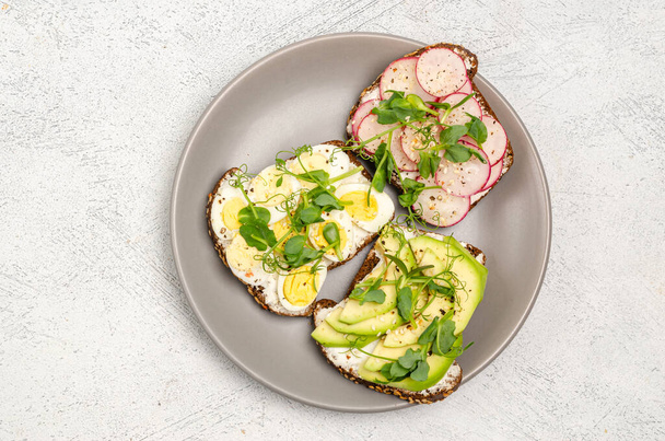 Different sandwiches with radishes, avacado, egg and microgreens on a plate on a light background. Flat lay, healthy snack. View from above. - Foto, immagini