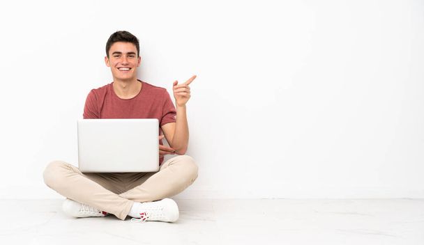Подросток сидит на флоре с ноутбуком, указывая пальцем в сторону. - Фото, изображение