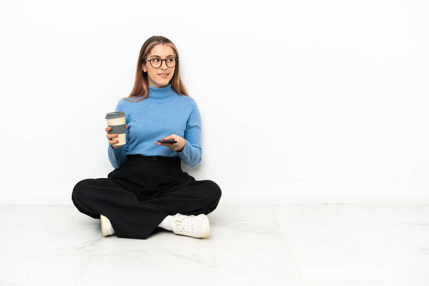 Jeune femme caucasienne assise sur le sol tenant du café à emporter et un mobile tout en pensant à quelque chose - Photo, image