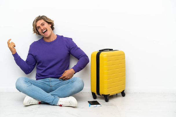 Young blonde man with suitcase sitting on the floor making guitar gesture - Photo, Image