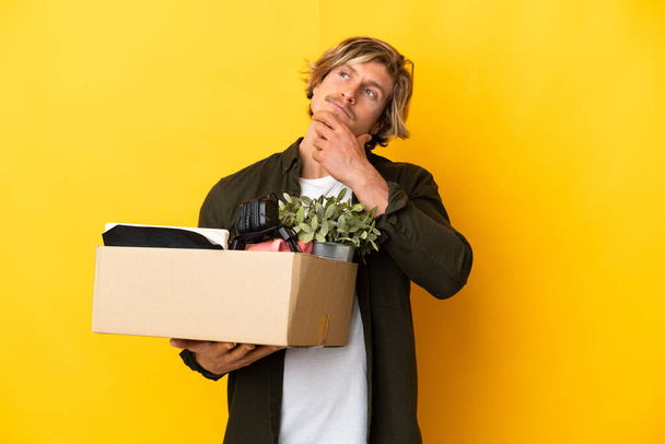 blonde man making a move while picking up a box full of things isolated on yellow background having doubts - Photo, Image