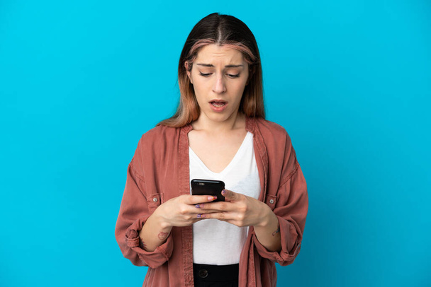 Young caucasian woman isolated on blue background looking at the camera while using the mobile with surprised expression - Photo, Image