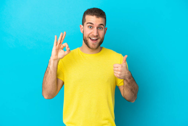 Молодий красивий кавказький чоловік ізольований на синьому фоні, показуючи знак ок і великий палець вгору жест
 - Фото, зображення