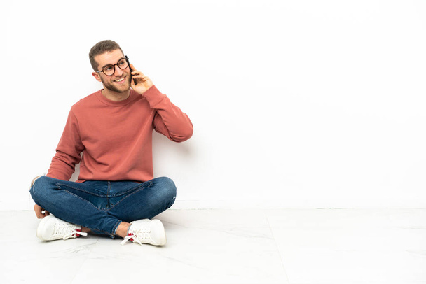Nuori komea mies istuu lattialla pitämässä keskustelua matkapuhelimen kanssa jonkun kanssa - Valokuva, kuva