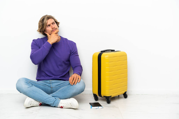Joven hombre rubio con maleta sentada en el suelo teniendo dudas y con expresión de cara confusa - Foto, Imagen