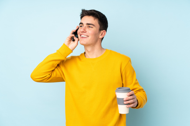 Έφηβος καυκάσιος όμορφος άντρας απομονωμένος σε μωβ φόντο κρατώντας καφέ για να τον πάρει και ένα κινητό - Φωτογραφία, εικόνα
