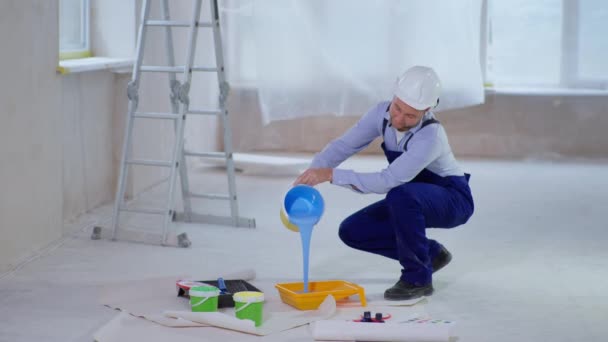 человек в рабочей одежде и кепке заливает голубую краску в поднос для покраски стен во время ремонта - Кадры, видео