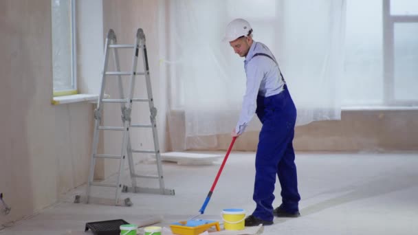 joven trabajando un trabajador de la construcción en traje de trabajo y casco con rodillo de construcción y cubo de pintura para reparar paredes - Metraje, vídeo