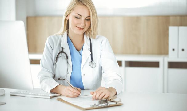 Woman-doctor at work while sitting at the desk in hospital or clinic. Blonde cheerful physician filling up medication history record form - 写真・画像