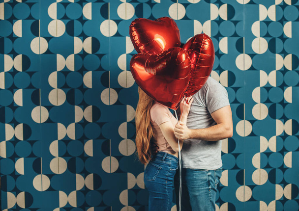 Kissing caucasian couple are hiding behind some air heart sharped balloons on a blue studio wall - 写真・画像