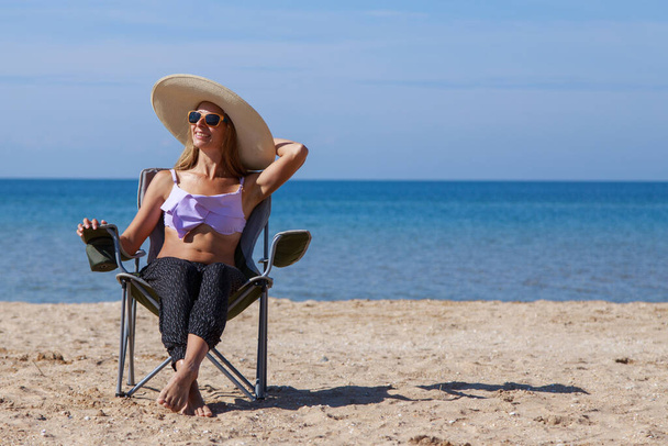 journey to the sea. girl in a bathing suit and hat sunbathing on the beach. tourist sitting on the sand. leisure wear. copy space - Photo, Image