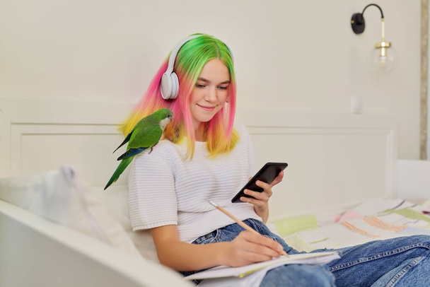 Girl student teenager sitting at home on bed with pet parrot on shoulder - Фото, изображение