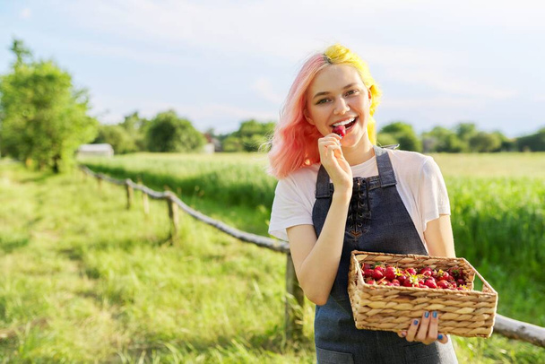 Beautiful smiling teenage girl on rustic farm with basket of fresh strawberries, copy space on green grass - Photo, image