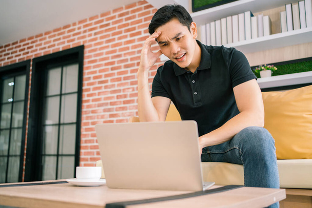 Asian male in casual feels stress and concerned with laptop while working and sitting on the sofa. Businessman working with problems on a laptop at home. Solving the problem process in business. - Foto, Imagen