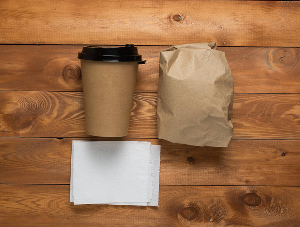 xícara de papelão com café e sanduíche embrulhado em papel. guardanapo branco. fundo de madeira. espaço de cópia. deitado plano. - Foto, Imagem