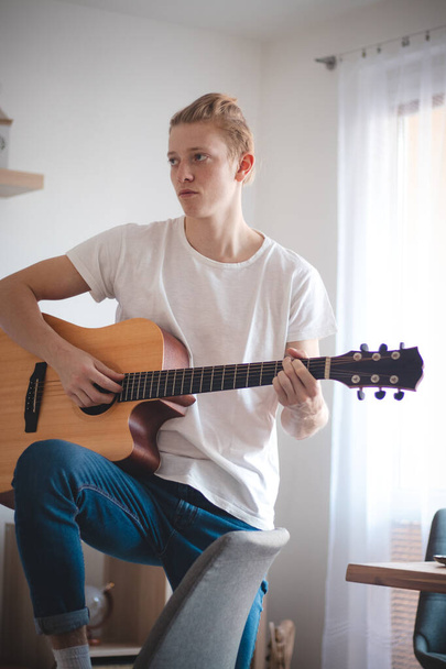 Candid portrait of a young artist in the living room. The singer holds his guitar and strums a song. Home quarantine. Fulfilling your dream. In a white T-shirt and blue jeans. - Photo, Image