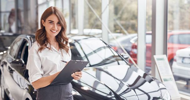 Positive saleswoman with clipboard working in car dealership - Zdjęcie, obraz