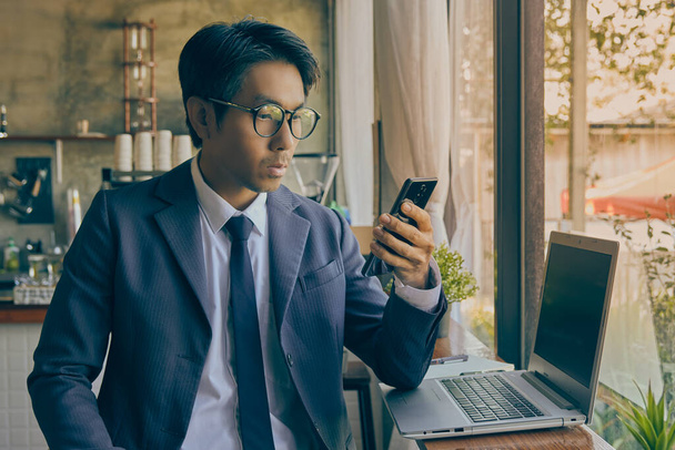 フロントライトアジアのビジネスマンがスーツを着て眼鏡をかけるコーヒーショップのシーンでスマートフォンやノートパソコンを使っています。アジアのビジネスマンはヴィンテージトーンの技術でどこからでも仕事 - 写真・画像