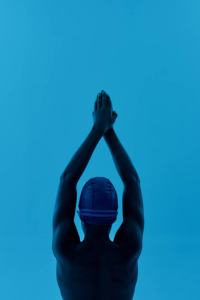 Young swimmer. Vertical shot of a teenage african boy in swimming cap standing in starting pose isolated over blue background, ready to dive - Photo, Image