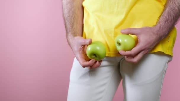  Der Mensch hält die Äpfel in seinen Händen auf Höhe der Genitalien und untersucht sie, indem er sie in verschiedene Richtungen dreht. Nahaufnahme. Rosa Hintergrund. Das Konzept der Männergesundheit - Filmmaterial, Video